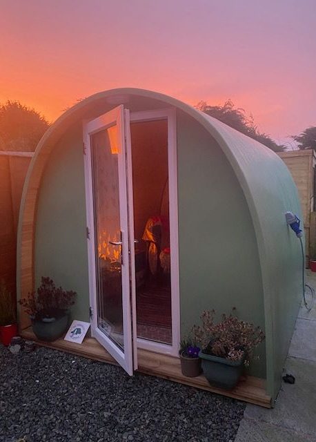 Green pod-shaped structure with a small wooden porch and open door at sunrise.