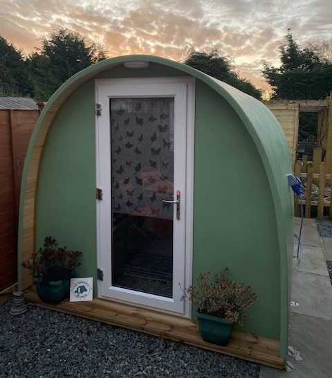 A green pod-shaped structure with a wooden frame, featuring a glass door and potted plants.