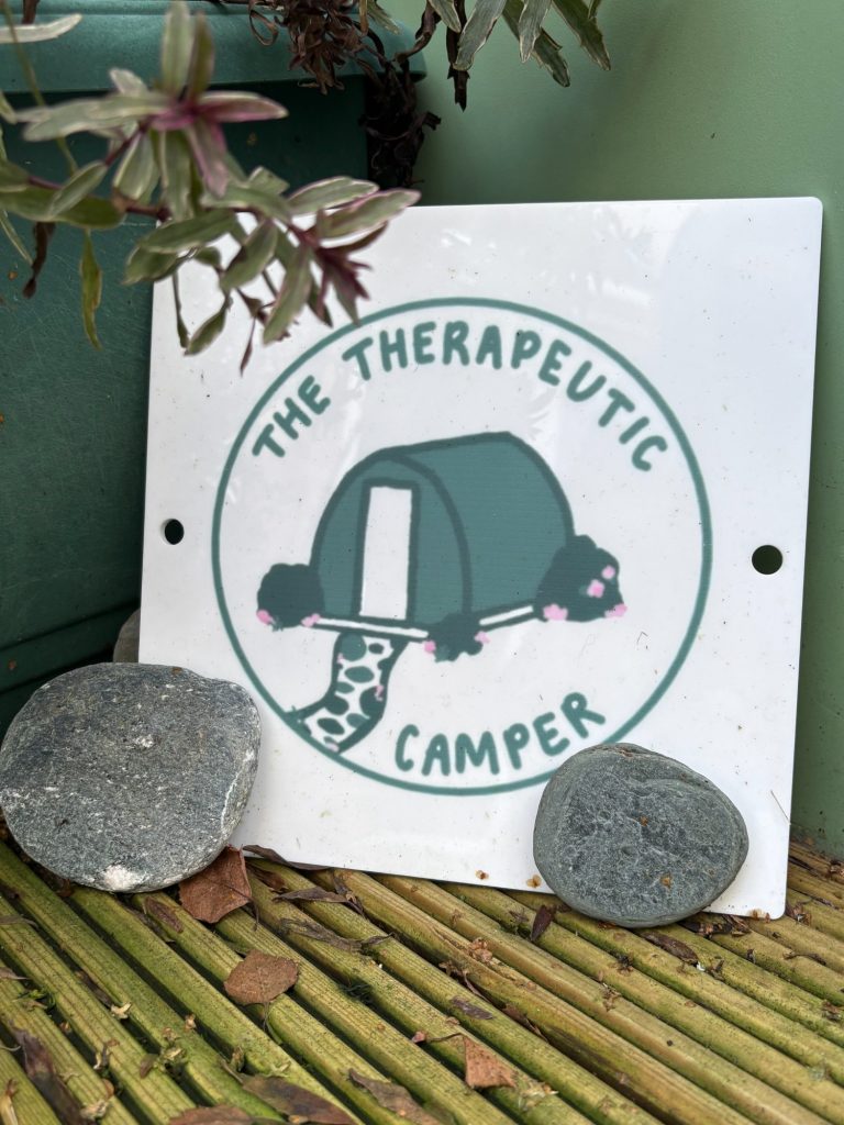 Logo of "The Therapeutic Camper" featuring a tent design on a sign with two stones.