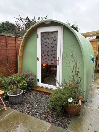 Green pod-shaped garden shed with a door and plants around it.