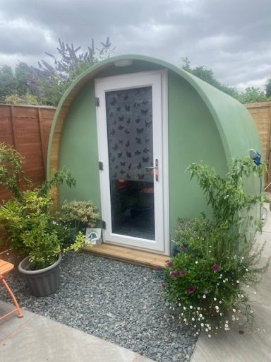 Small green pod-shaped cabin surrounded by plants and gravel, with a wooden entrance.