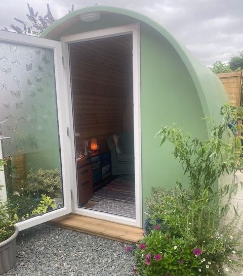 The Therapeutic Camper Pod A green pod-shaped structure with a wooden frame, featuring a glass door and potted plants.